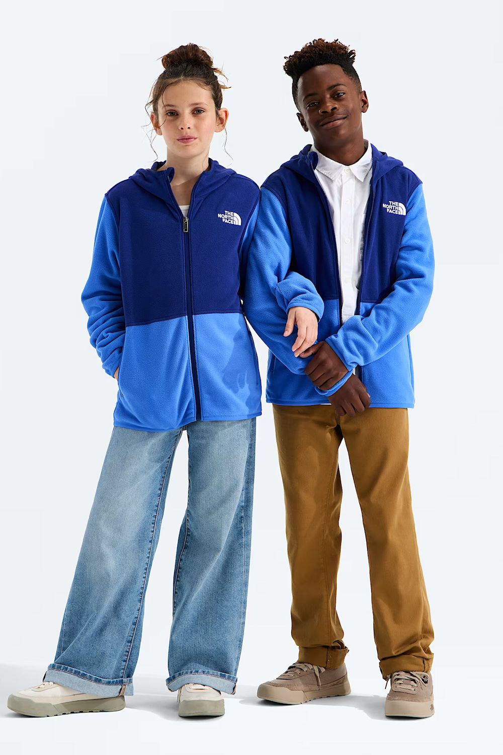 Two models wearing blue and royal blue colorblock zip-up hoodies by The North Face, styled with jeans and brown pants against a light gray studio background.