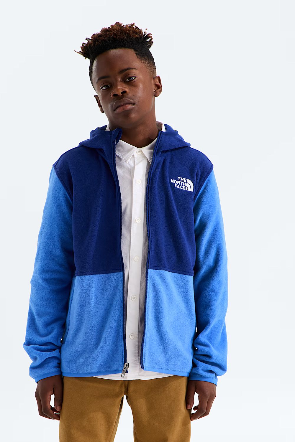 A young man wearing a blue and navy fleece zip-up hoodie by The North Face, layered over a white collared shirt and tan pants, set against a plain light gray studio background.