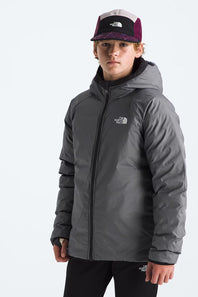 A boy wearing a dark gray The North Face hooded puffer jacket and a black, white, and purple trucker hat, standing against a light gray studio background.