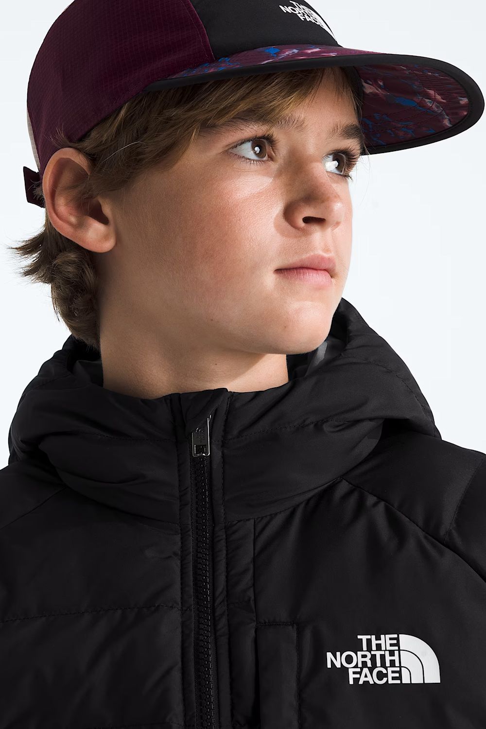 A close-up of a child wearing a black puffer jacket with a white The North Face logo on the chest and a dark maroon and black trucker cap, photographed against a light gray studio background.