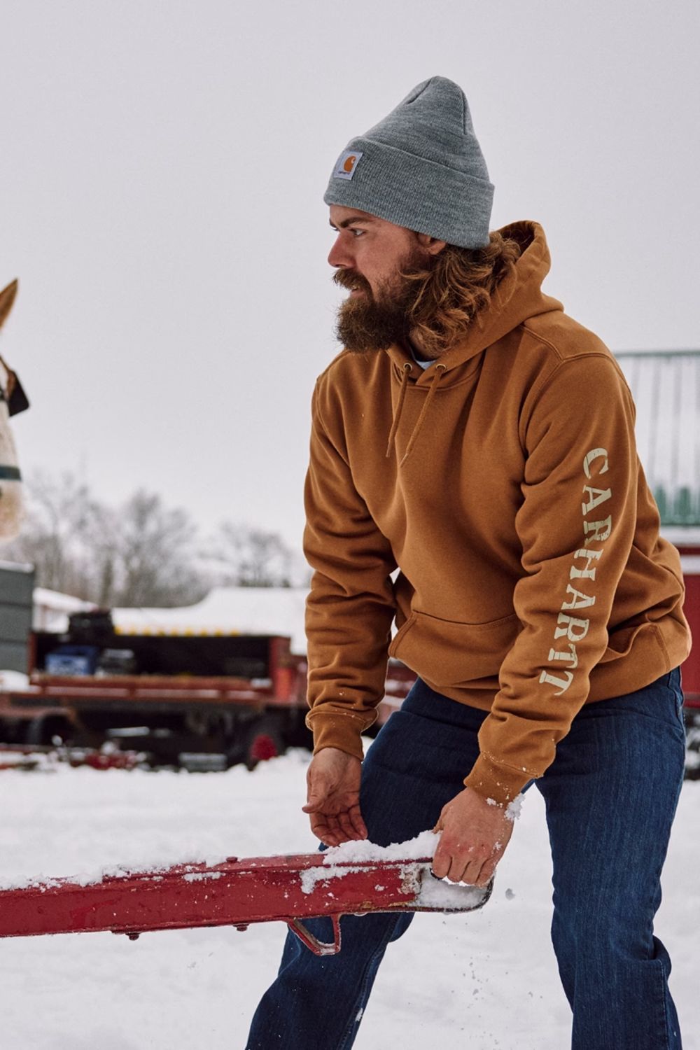 A man wearing a brown Carhartt hoodie and blue jeans stands in a snowy outdoor setting next to a horse-drawn sled, with the Carhartt brand name visible on the sleeve of the hoodie.