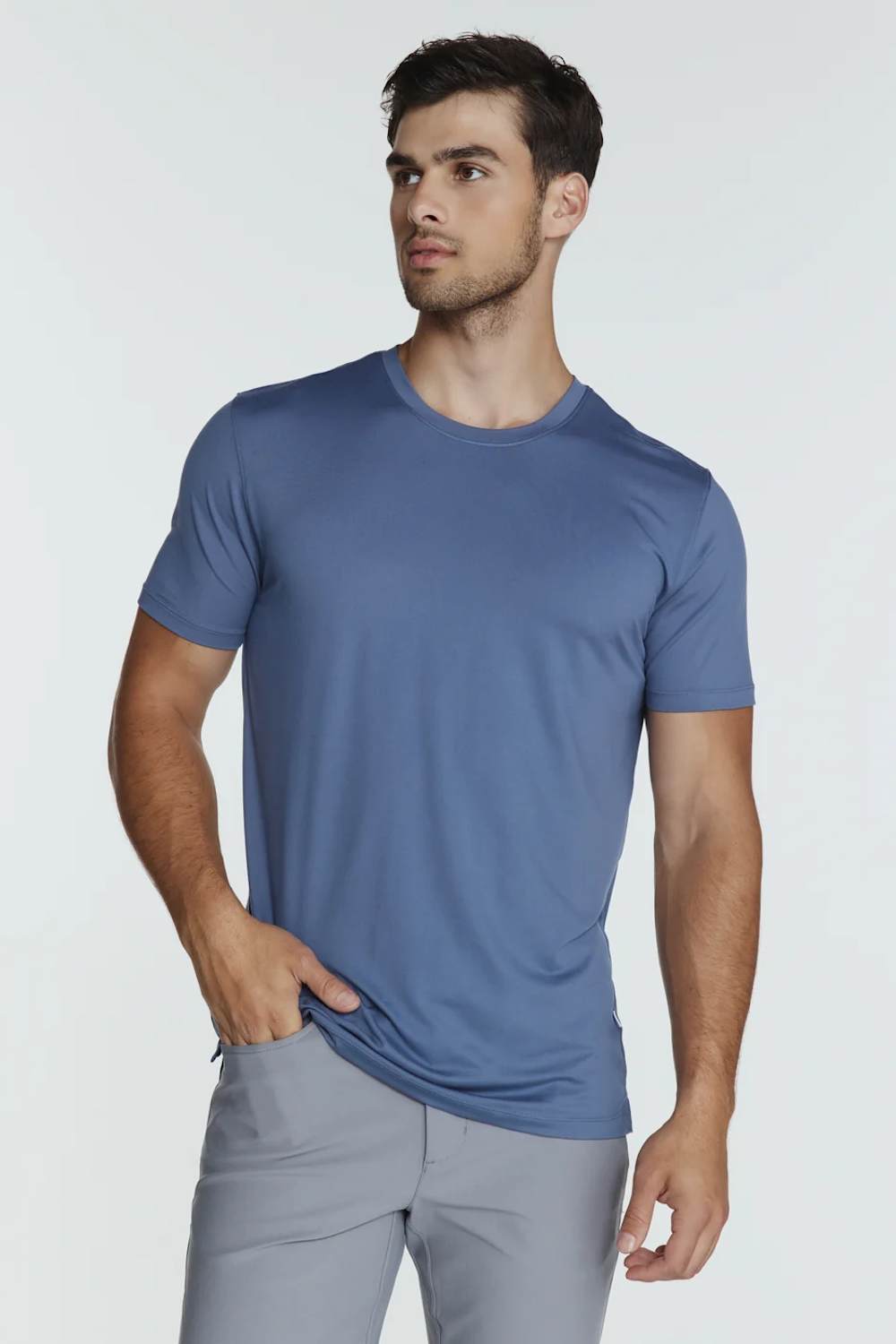 A man wearing a slate blue crew-neck t-shirt and light gray trousers, standing against a light gray studio background.