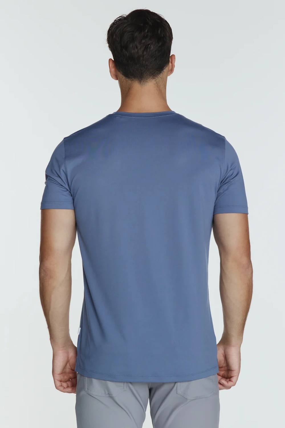A man viewed from behind wearing a slate blue short-sleeve athletic t-shirt and light gray pants, set against a plain light gray studio background.