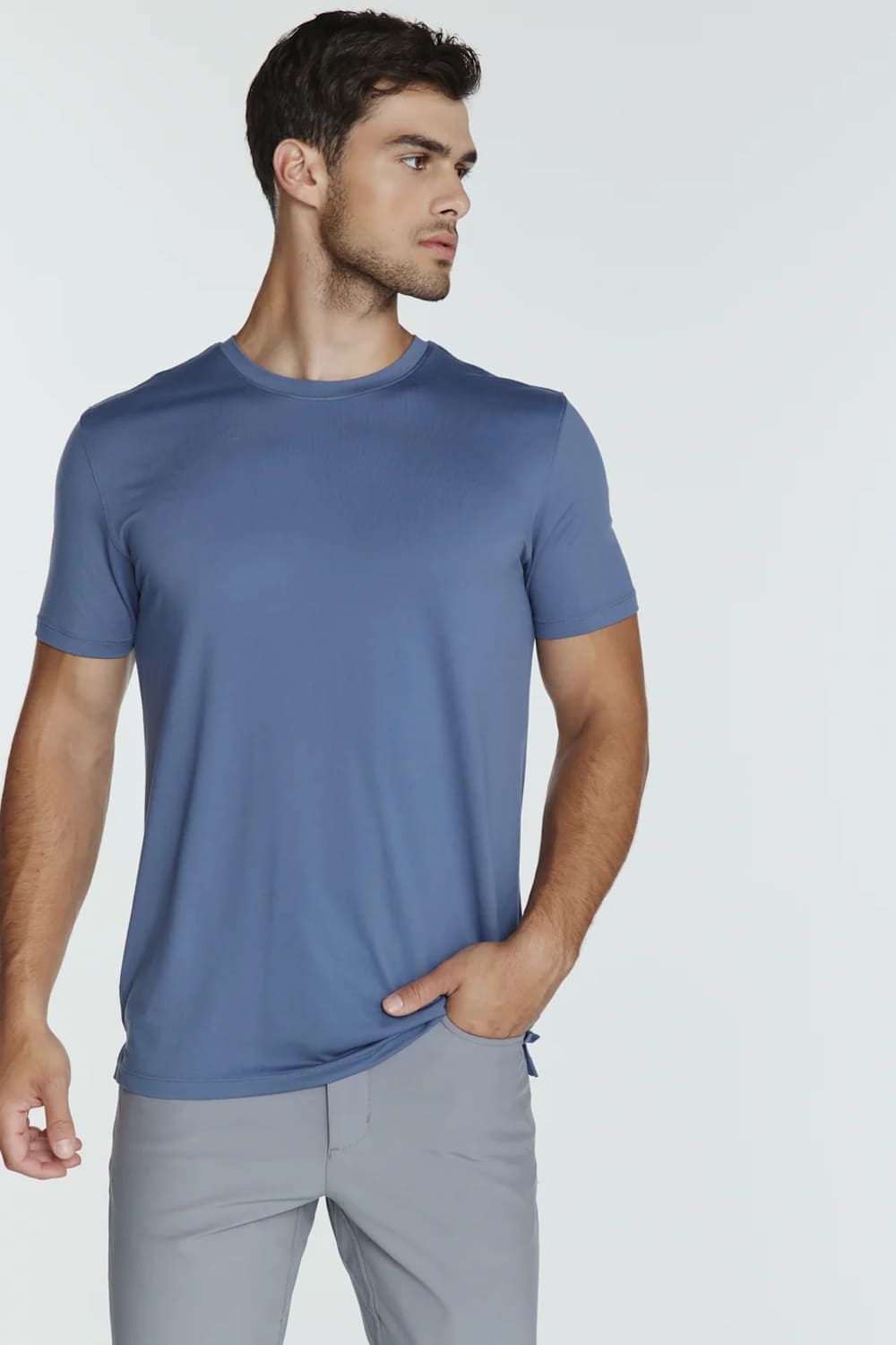 A man wearing a slate blue short-sleeve crewneck t-shirt and light gray trousers, standing against a light gray studio background.