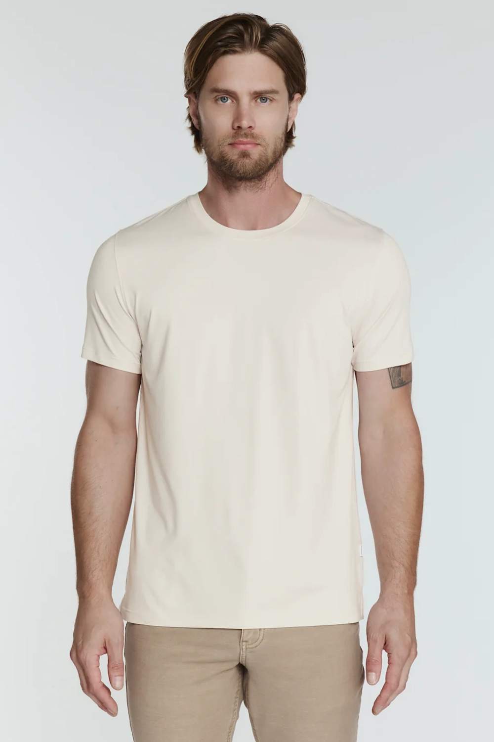 A man wearing a plain cream-colored short-sleeve crewneck t-shirt and khaki pants, standing against a light gray studio background.