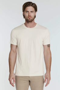 A man wearing a plain cream-colored short-sleeve crewneck t-shirt and khaki pants, standing against a light gray studio background.