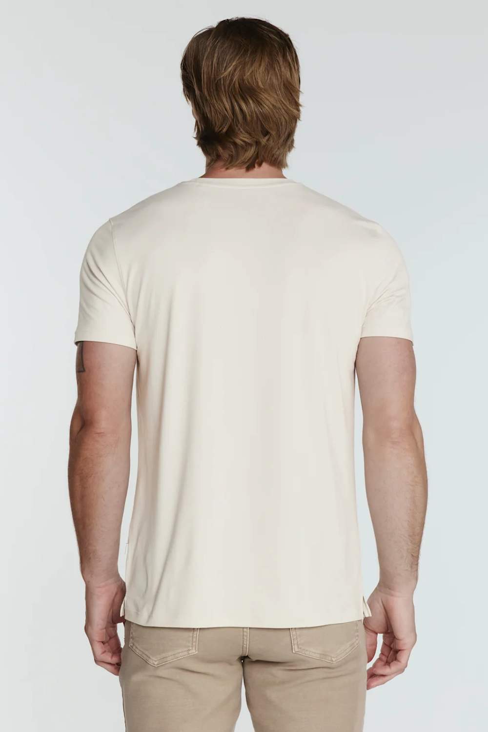 A man viewed from behind wearing a plain cream-colored short-sleeve t-shirt and khaki pants, set against a light gray studio background.