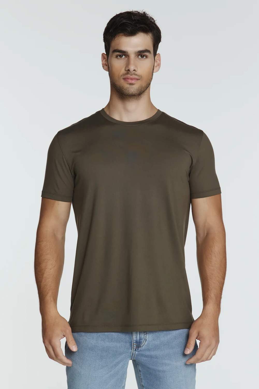 A man wearing a short-sleeve crewneck t-shirt in olive brown, paired with light blue jeans, standing against a light gray studio background.