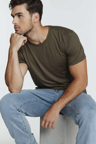 A man wearing an olive green crew-neck t-shirt and light blue jeans, sitting on a light gray block against a plain light gray background.