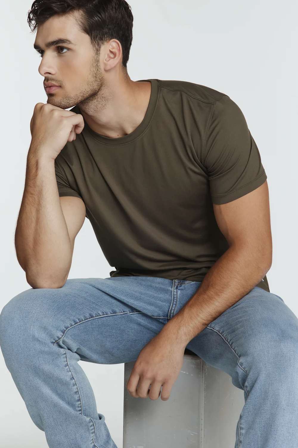A man wearing an olive green crew-neck t-shirt and light blue jeans, sitting on a light gray block against a plain light gray background.