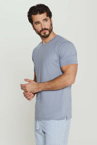 A man wearing a light blue-gray short-sleeve crewneck t-shirt and light blue patterned pajama pants, standing against a plain light gray studio background.