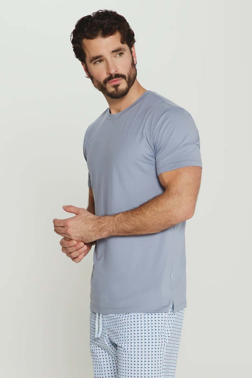 A man wearing a light blue-gray short-sleeve crewneck t-shirt and light blue patterned pajama pants, standing against a plain light gray studio background.