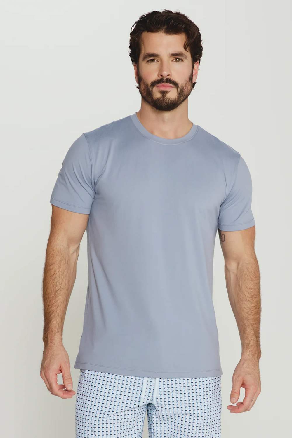A man wearing a light blue-gray crewneck t-shirt and white patterned shorts, standing against a light gray studio background.