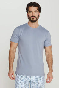 A man wearing a light blue-gray crewneck t-shirt and white patterned shorts, standing against a light gray studio background.