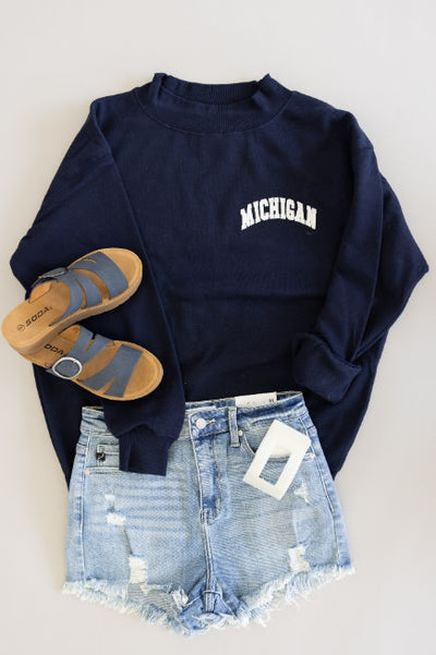 Women's flat lay outfit featuring light wash distressed denim shorts, a mock neck navy sweatshirt with 