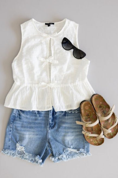 Women's flat lay outfit including medium wash denim shorts, a white peplum style top with bow details, black sunglasses, and Birkenstock sandals