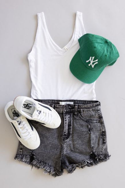 Women's outfit flatlay featuring black acid wash denim shorts, a white tank tank, white Vans sneakers, and kelly green New York Yankees hat