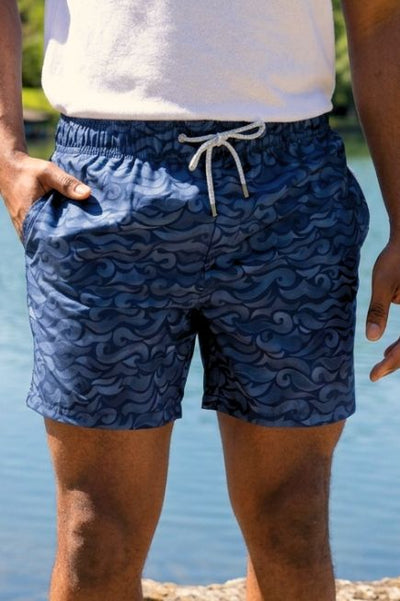 Man wearing blue/black ocean wave printed swim trunks and a white t-shirts standing in front of a lake