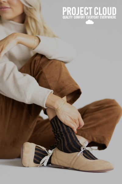 Promotional image for Project Cloud featuring a woman sitting seated on a white background. She is wearing tan suede ballet flats with white laces tied in a bow, styled over black and brown striped socks, brown corduroy pants, and a cream-colored sweater.