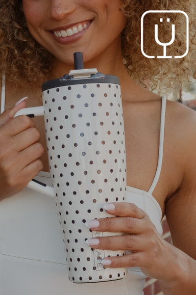 Close-up of a smiling curly-haired woman holding a white BrüMate insulated tumbler with a black polka dot pattern, white side handle, and built-in straw.