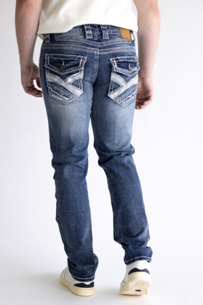 Back view of a person modeling blue embellished jeans with white stitching, a white top, and sneakers against a plain background.