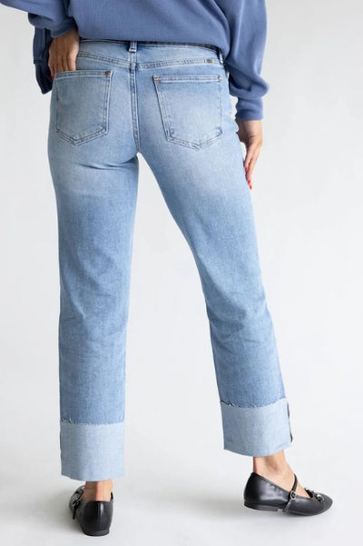Rear view of a model wearing light-wash cuffed jeans, a blue sweater, and black loafers in a minimalist studio setting.