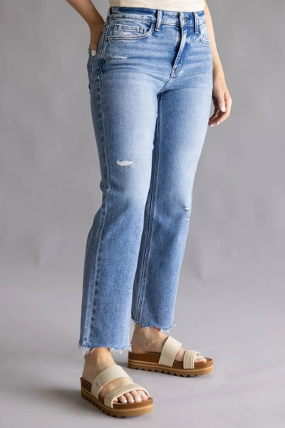 A person in light blue, slightly distressed jeans with raw hems and beige platform sandals stands against a gray background, conveying a casual and modern style.