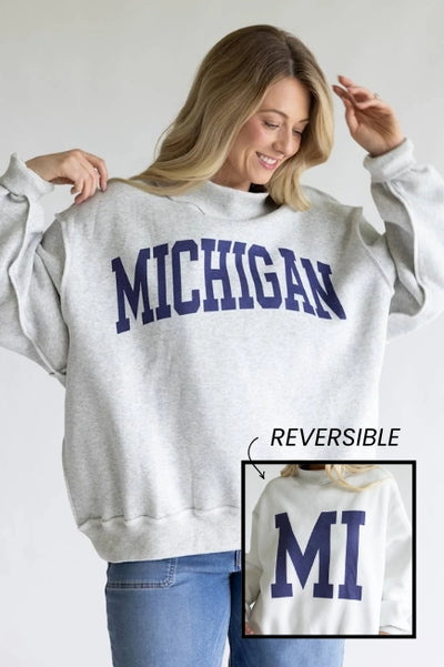 A woman modeling an oversized mock neck sweatshirt with MICHIGAN screen-printed. In the lower right corner, an insert photo labeled REVERSIBLE showing the inside of the sweatshirt with MI screen-screenprinted