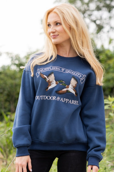 A woman outdoors, wearing a navy crewneck sweatshirt featuring mallard ducks with text Suppliers & Quality, Outdoor & Apparel paired with black jeans