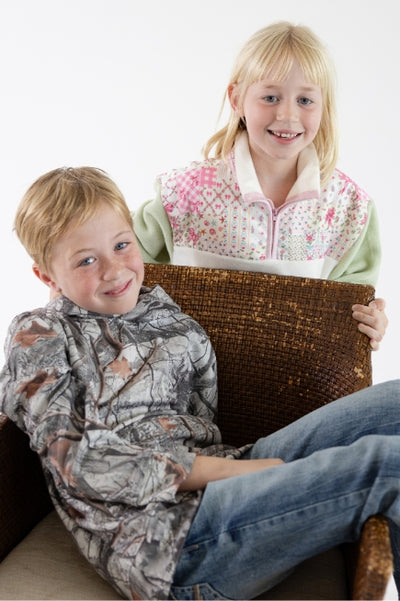 2 kids posing together; One kid is a boy sitting in a brown wicker cushioned chair wearing a camo hoodie and denim jeans. The other kids is a girl standing behind the wicker chair wearing a quilted patchwork type of quarter zip in front of a white background