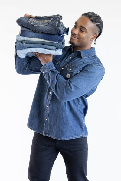 A man standing in front of a white background wearing a long sleeve button up denim shirt and black pants holding a stack of denim jeans