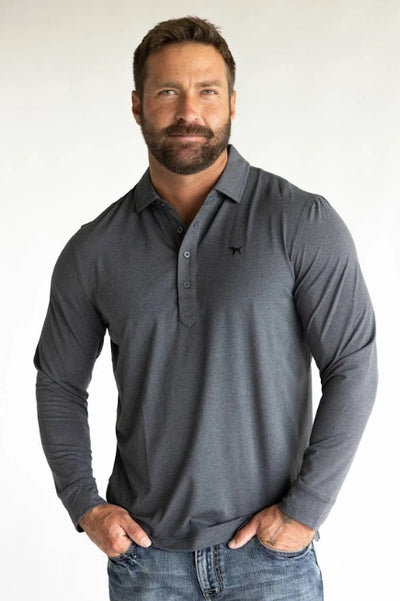 A man standing in front of a plain background wearing a grey long sleeve polo shirt with denim jeans