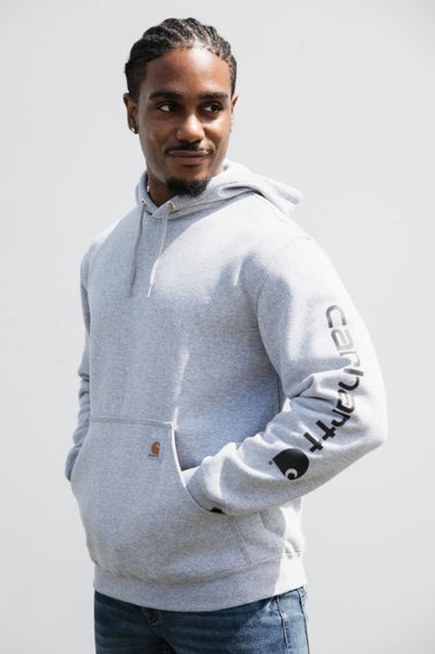 A man standing in front of a light grey background wearing a light great hoodie with Cahartt brand logo down the sleeve and denim jeans