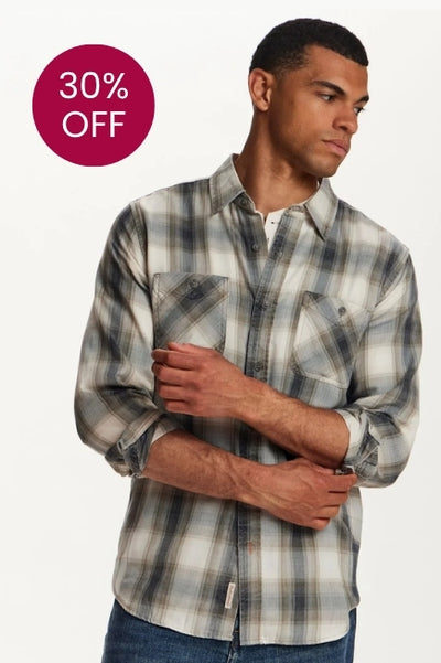 A man standing in front of a white background wearing a black, grey and beige flannel shirt and denim jeans. In the extra white space there is a maroon colored circle with 30% OFF