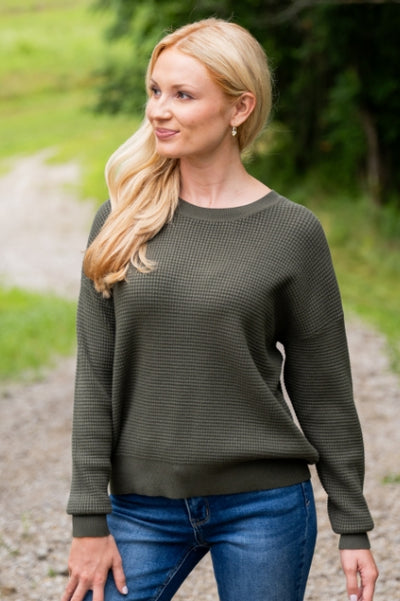 Woman standing outdoors on a gravel pathway wearing a dark olive green sweater and dark wash blue jeans