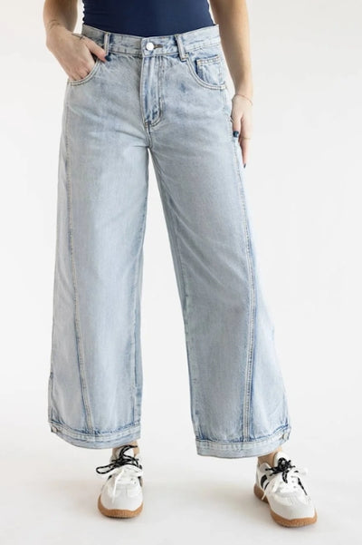 A person wearing light wash barrel leg jeans with a navy top and white sneakers standing in front of a white studio background