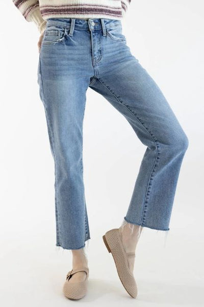 A woman wearing medium wash straight legs jeans with her knee popped up with a white and purple striped sweater and beige Mary Jane shoes on a white background