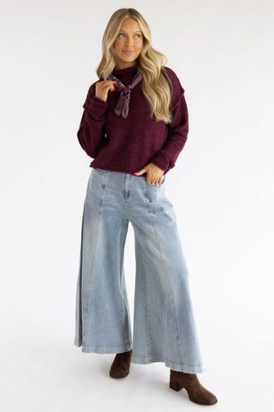 A woman wearing a maroon colored wearing with light wash palazzo styled jeans paired with an ascot and brown boots standing in front of a basic studio background