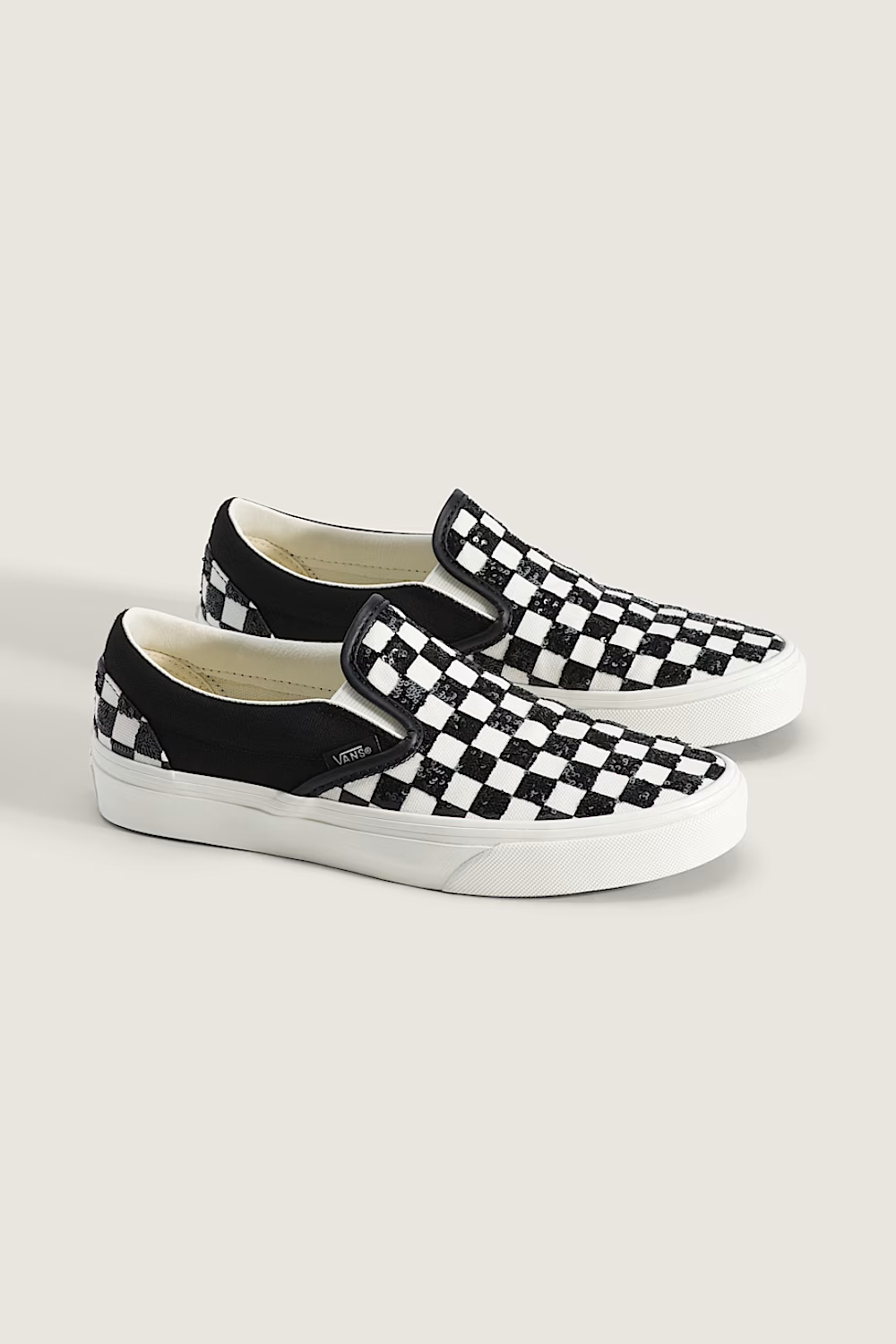 A pair of slip-on platform sneakers featuring a black and white checkerboard pattern on the uppers, with black canvas side panels and white rubber soles, shown against a light beige background.