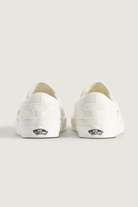 A pair of white Vans slip-on sneakers shown from the back, featuring a checkerboard pattern on the heel and the classic black Vans side logo on a light beige background.