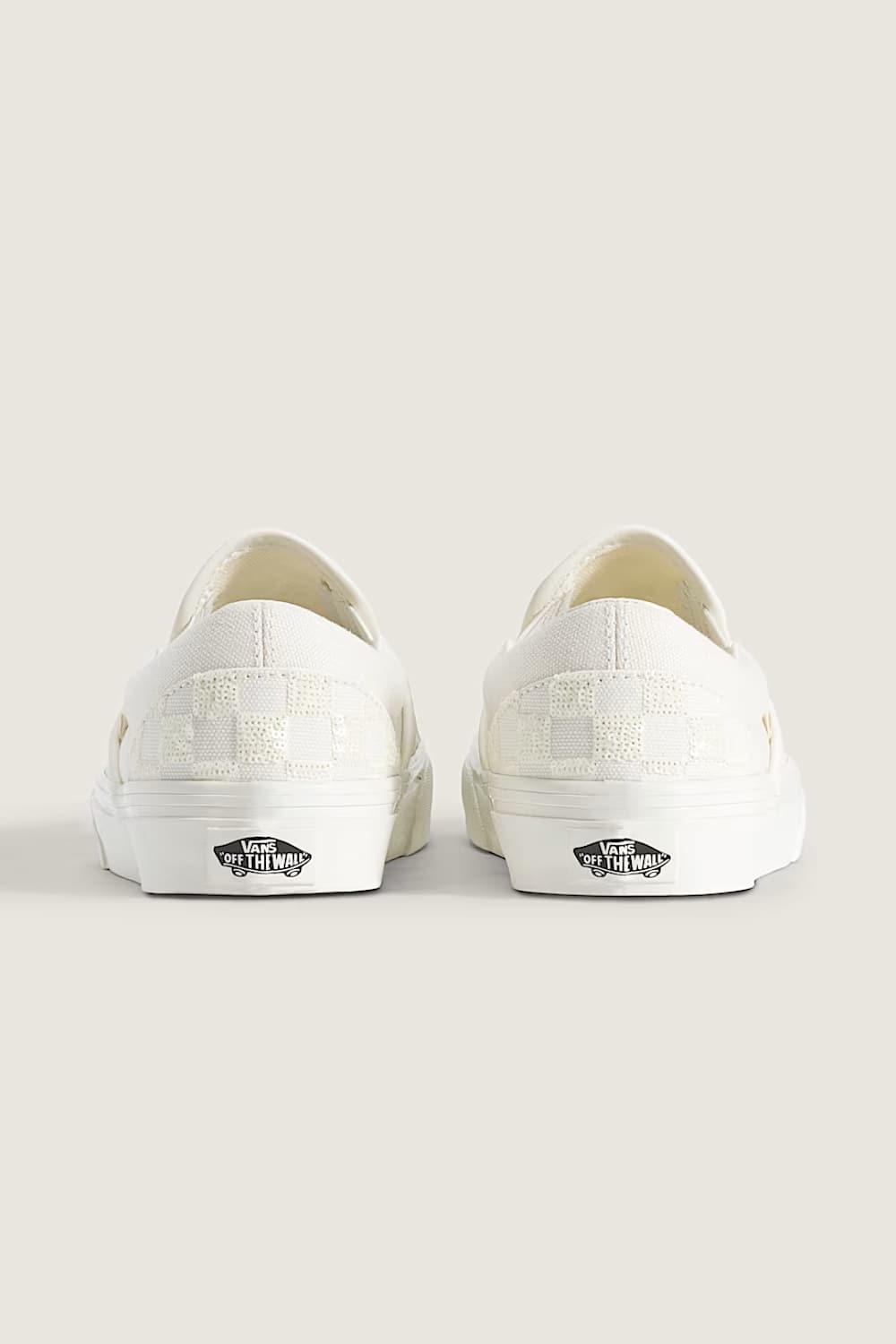 A pair of white Vans slip-on sneakers shown from the back, featuring a checkerboard pattern on the heel and the classic black Vans side logo on a light beige background.