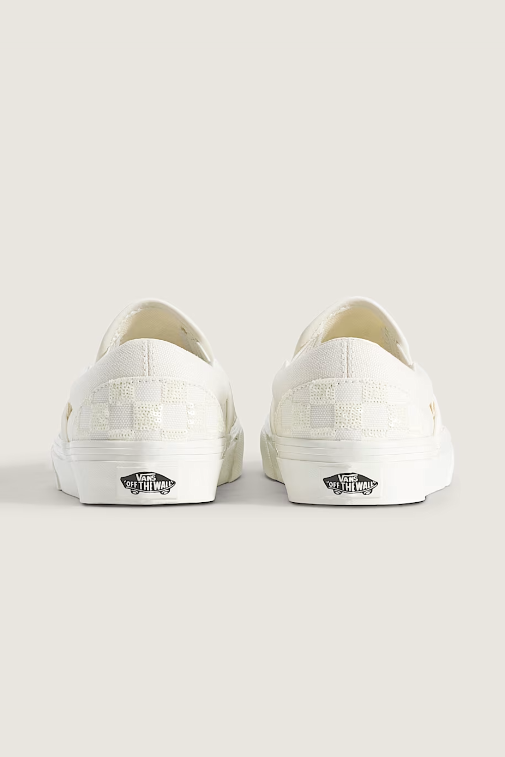 A pair of white Vans slip-on sneakers shown from the back, featuring a checkerboard pattern on the heel and the classic black Vans side logo on a light beige background.
