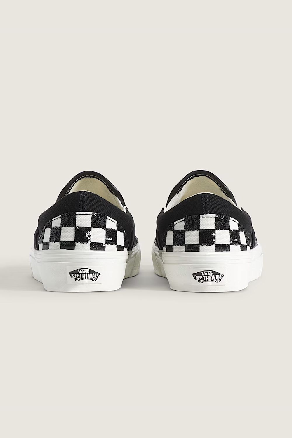 A pair of Vans slip-on sneakers shown from the back, featuring a black and white checkerboard pattern on the sides and white rubber soles with the Vans logo.