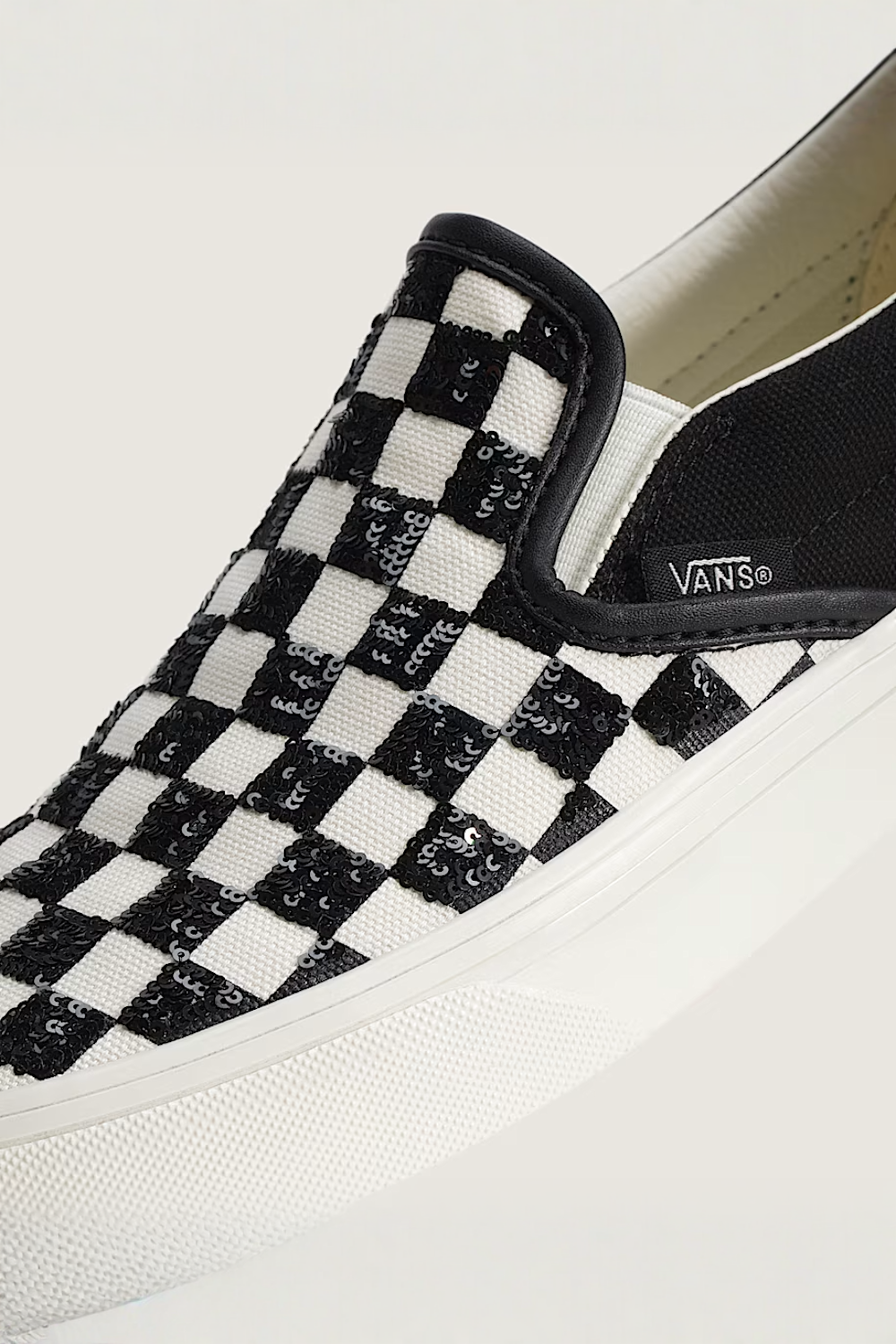 A close-up of a Vans slip-on shoe featuring a black and white checkerboard pattern with sequined details, a black heel tab, and a white rubber sole.
