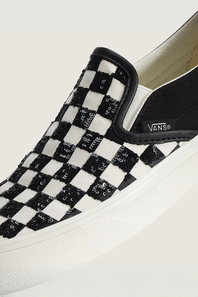 A close-up of a Vans slip-on shoe featuring a black and white checkerboard pattern with sequined details, a black heel tab, and a white rubber sole.