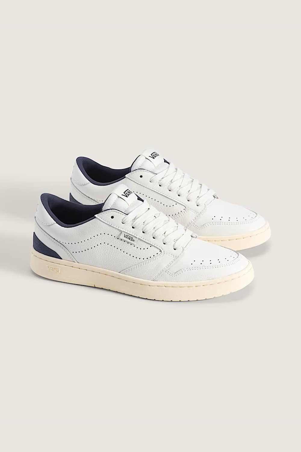 A pair of white leather low-top sneakers with dark navy blue accents on the collar lining and heel tab, set against a solid light beige background.