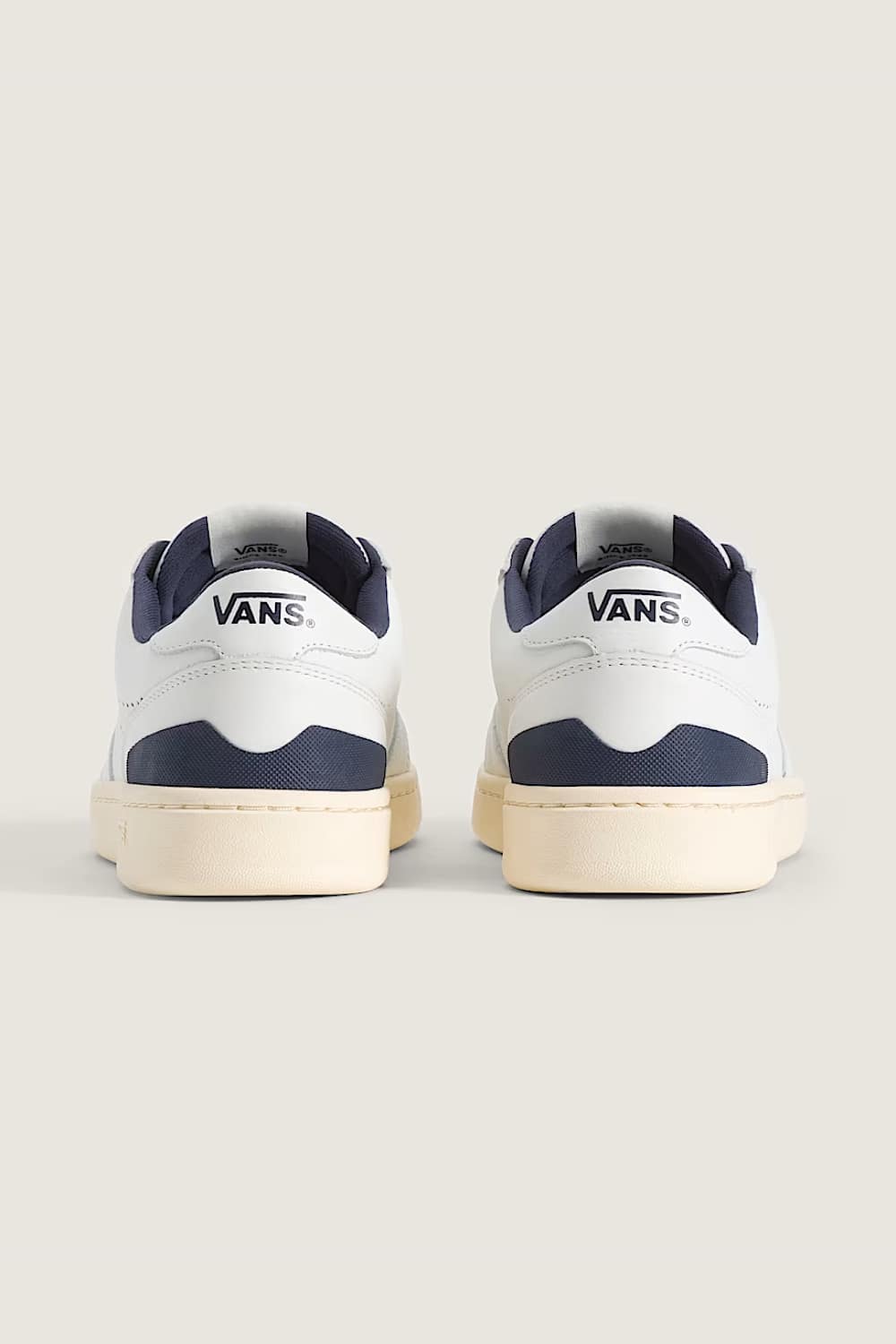 A pair of Vans low-top sneakers shown from the back, featuring a white leather upper with navy blue accents and a cream-colored sole, set against a solid light beige background.