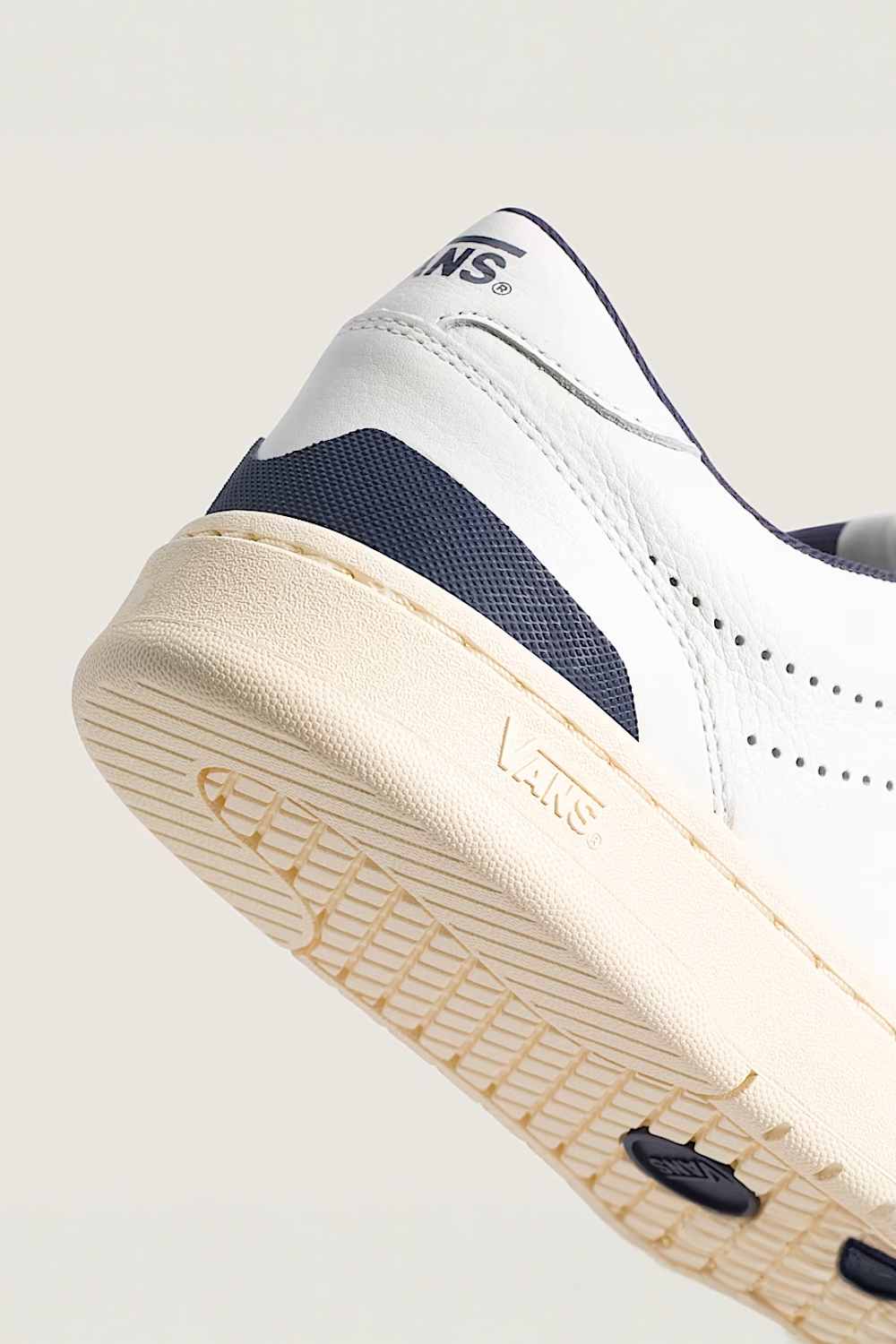 Close-up of a Vans sneaker showing the side and sole, featuring a white leather upper with navy blue accents and a cream-colored rubber sole.