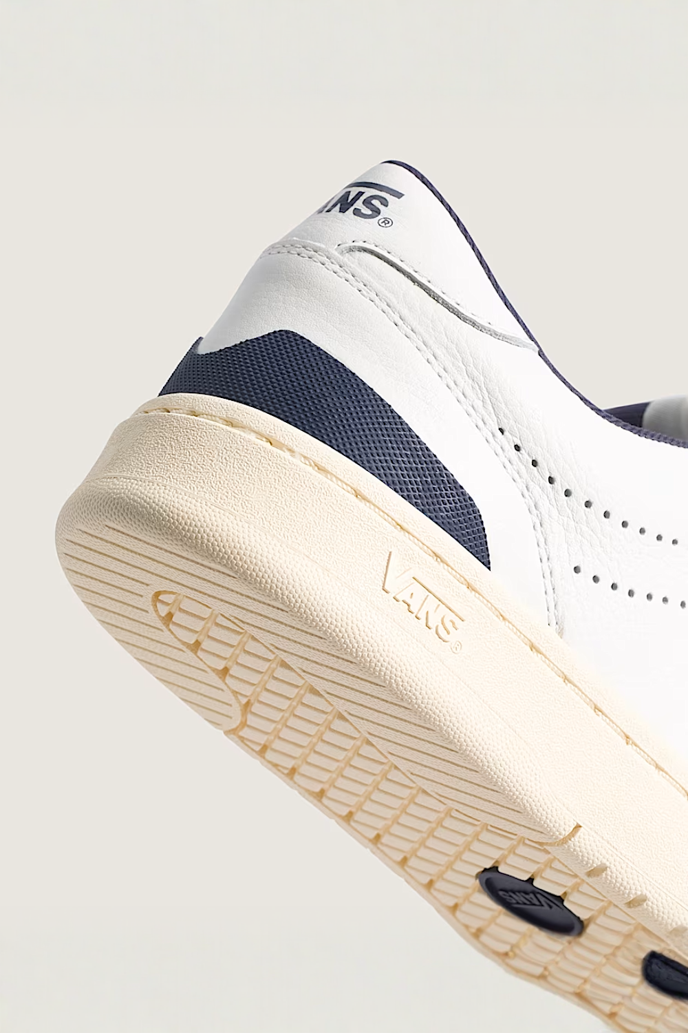 Close-up of a Vans sneaker showing the side and sole, featuring a white leather upper with navy blue accents and a cream-colored midsole.