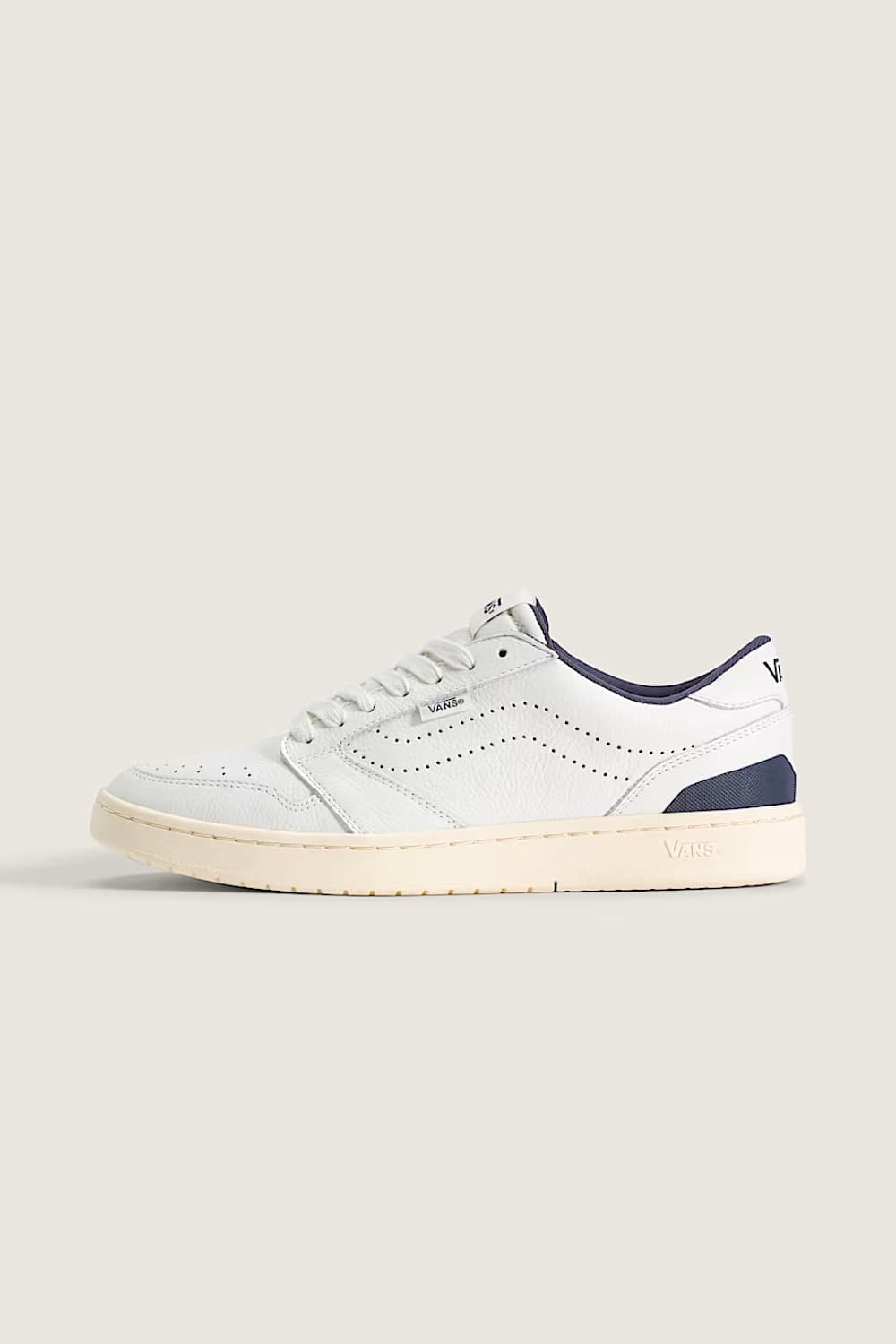 A low-top sneaker in white leather with navy blue accents on the collar, heel, and tongue, featuring a cream-colored rubber sole, shown against a solid light beige background.